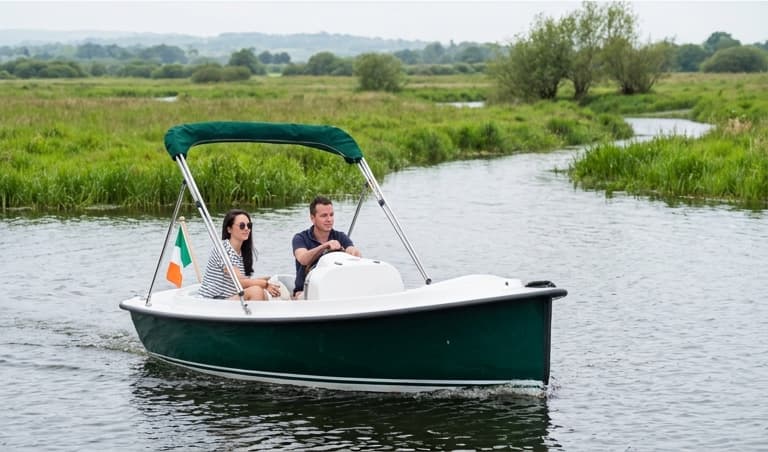 A couple aboard The Kingfisher on the Shannon Callows, one at the helm and one sitting alongside, meadow and open water behind