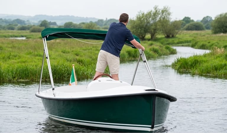 View from the water of a man steering The Kingfisher from the standing helm position, meadows either side